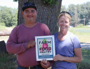 A man and woman holding a Farm to Food pantry placard outdoors, smiling at the camera