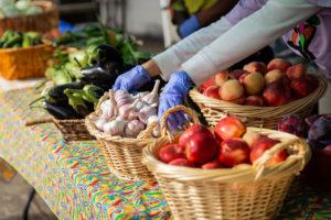 A person's gloved hands putting a wicker basket full of garlic bulbs onto a clothed table. Other baskets on the table are filled with apples, nectarines, plums, eggplants, and corn.