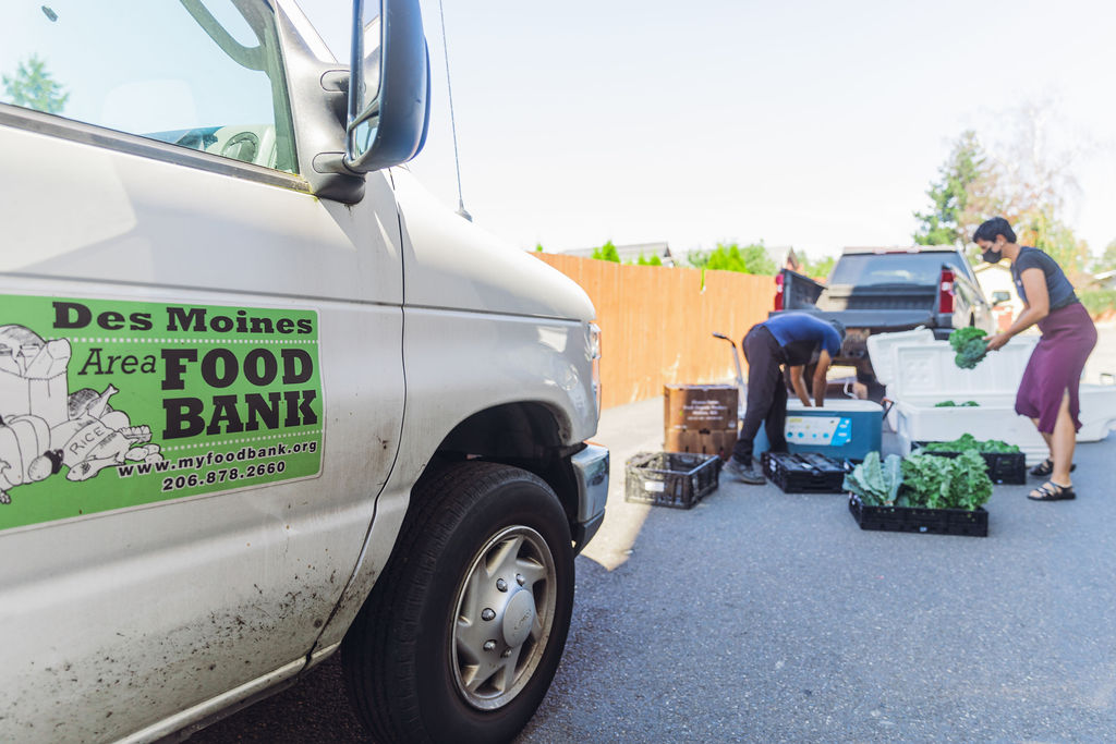 In the foreground, a white delivery van for the Des Moines Area Food Bank. In the background, a man and woman picking up crates of leafy green vegetables.